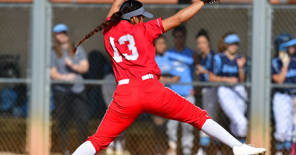 A softball player in a red uniform winding up to pitch on a dirt field as other players stand behind a fence.
